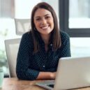 Shot of smiling business woman working with laptop while looking at camera in modern startup office.